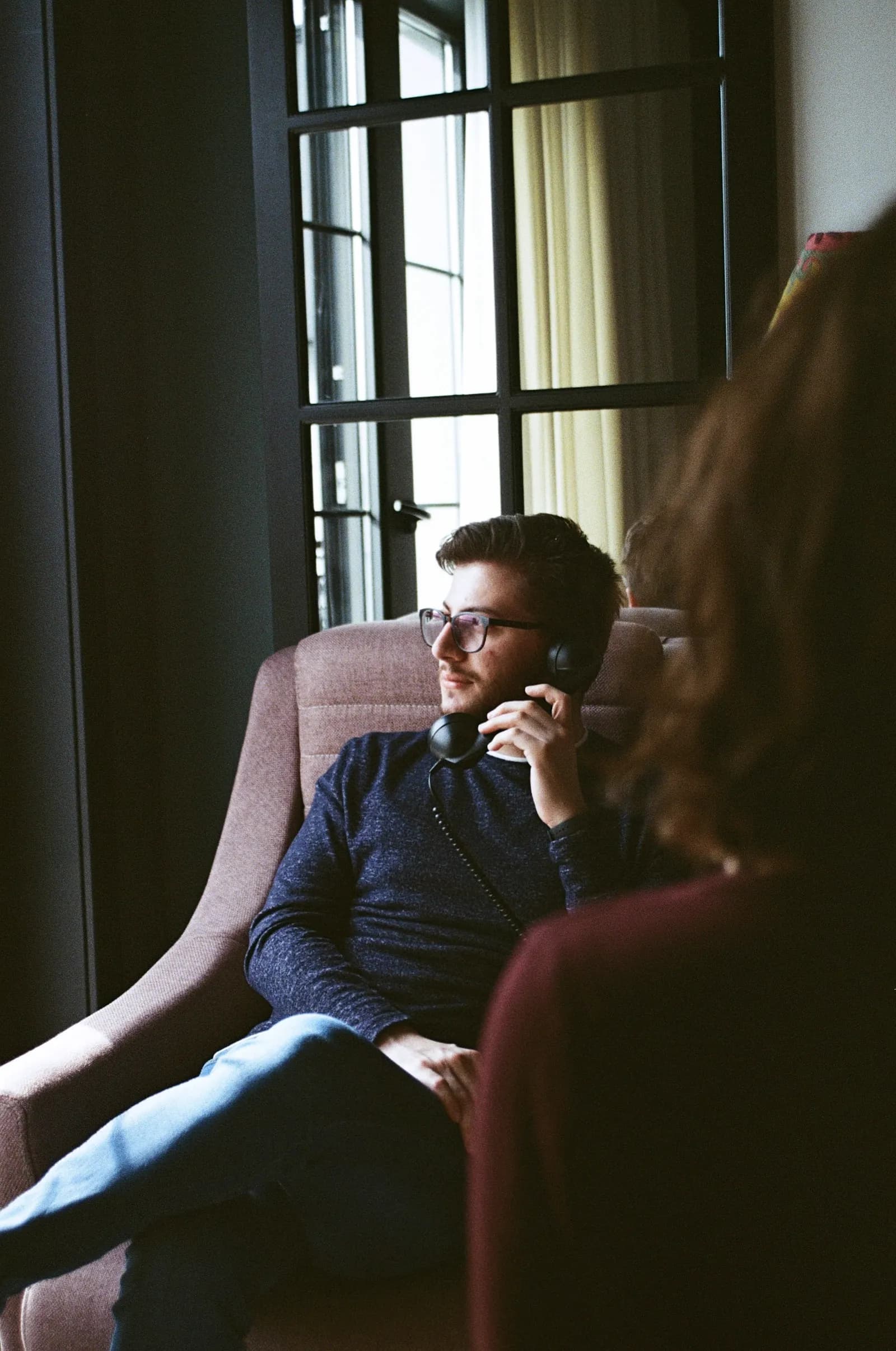 A man in an armchair by a window on a phone call.
